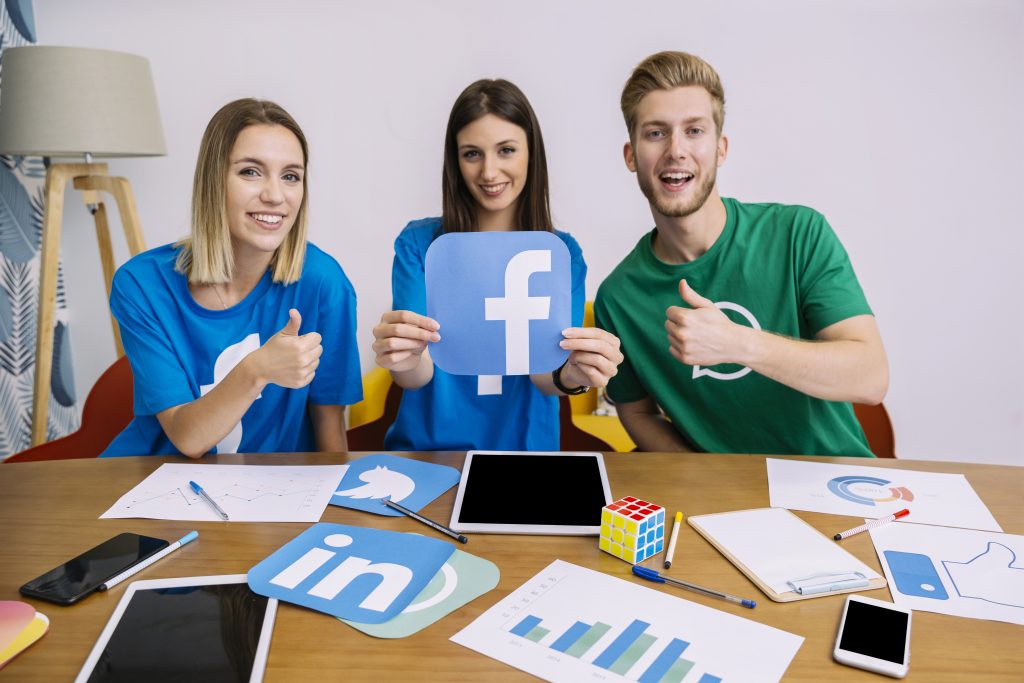 Three young professionals showing social media logos and giving thumbs up at a table with digital charts and mobile devices.
