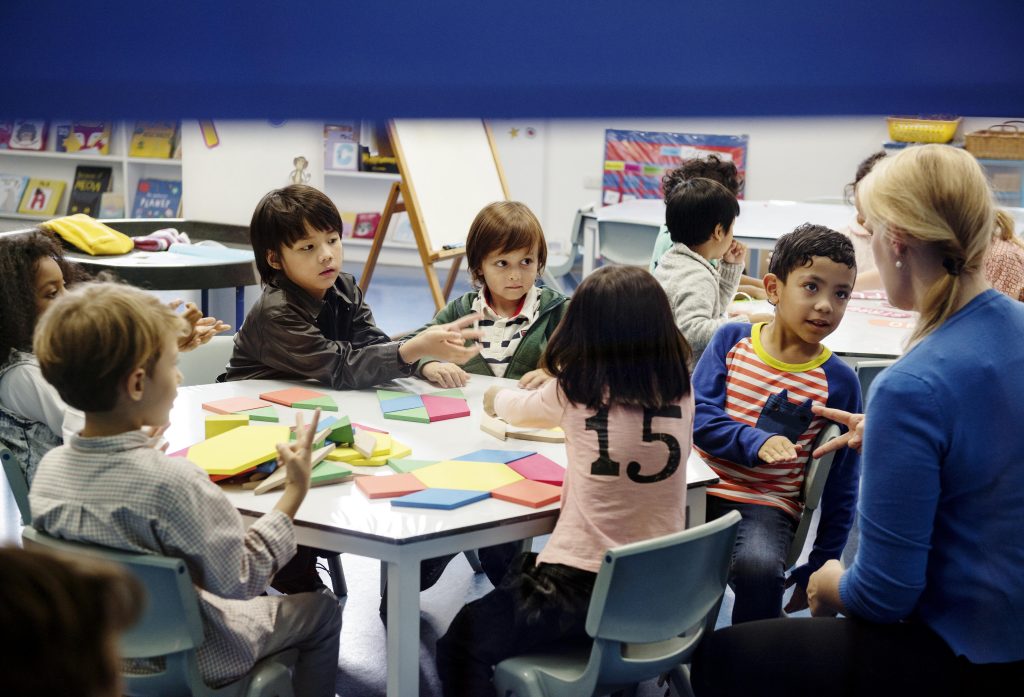 Young children sit around a table with colorful shapes, engaging in a classroom activity with a teacher in a vibrant setting.