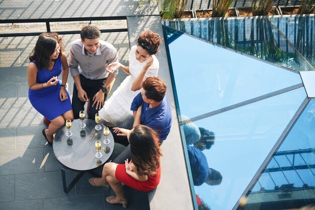 A group of five stylish young adults chatting and enjoying drinks around a small table on a rooftop patio near a glass structure.