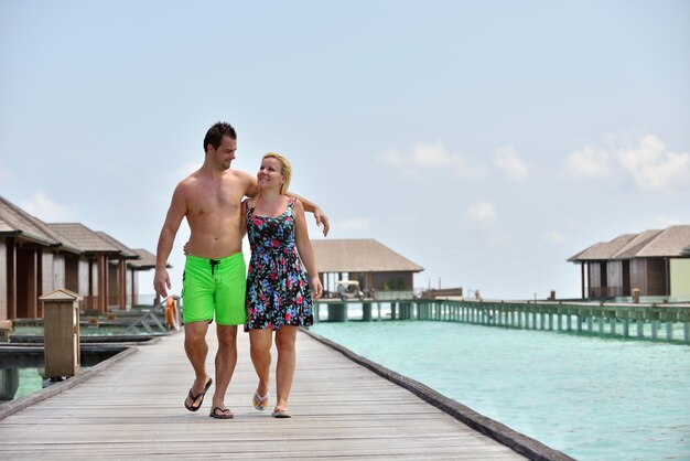 Happy couple walking arm in arm on a wooden pier over turquoise water, surrounded by overwater bungalows on a sunny tropical day.