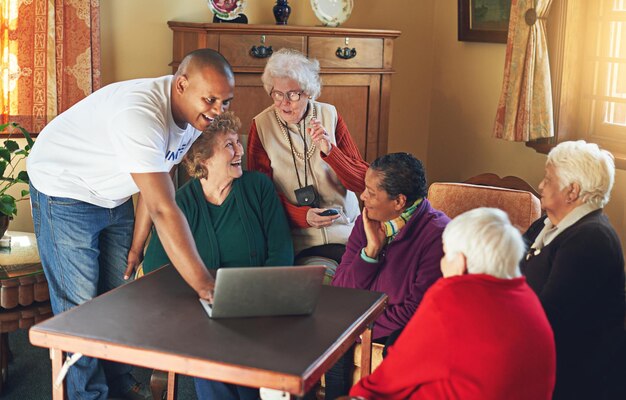 Retirees gather around a table as a caregiver shows a laptop, sharing smiles, conversation, and warm social moments indoors.