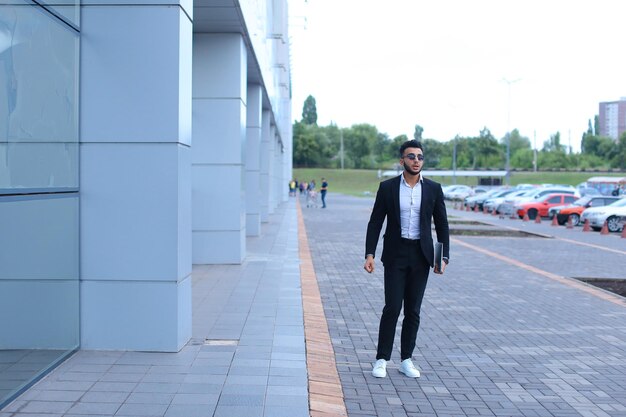 Man in a black suit and white sneakers walking outside a modern building, holding a laptop, with parked cars and greenery behind him.