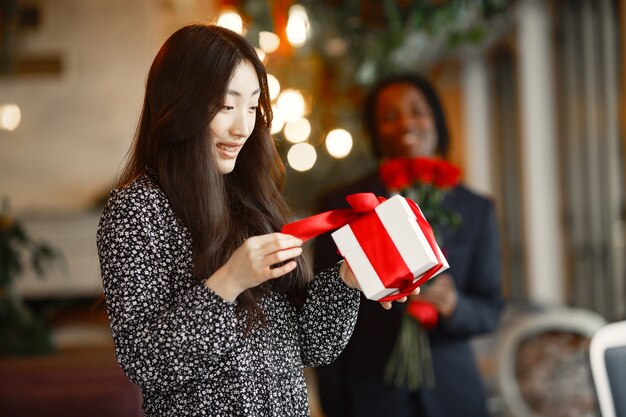 Dark-skinned man giving roses to a happy woman with a gift at a café.