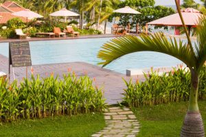 Tropical resort poolside with sun loungers, umbrellas, palm trees, a winding garden path, and serene, blue ocean waters nearby.