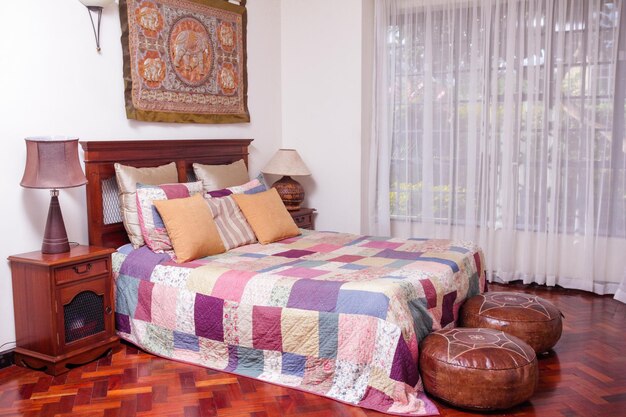 A bright bedroom with a colorful patchwork quilt, wooden furniture, sheer curtains, and two round leather ottomans at the foot of the bed.
