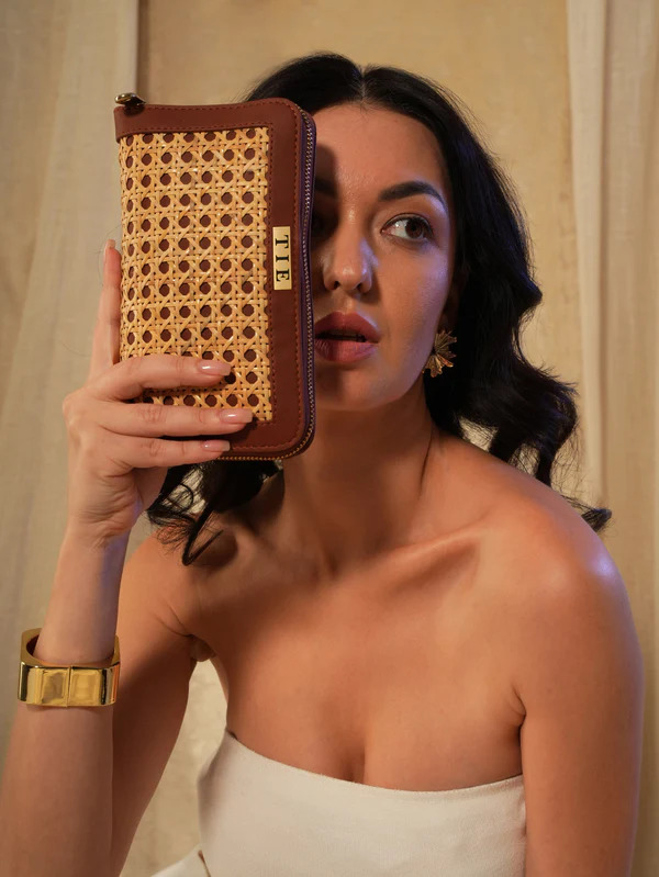 A woman in a white strapless top holds a woven rattan and brown leather clutch partially covering her face, accessorized with gold jewelry.